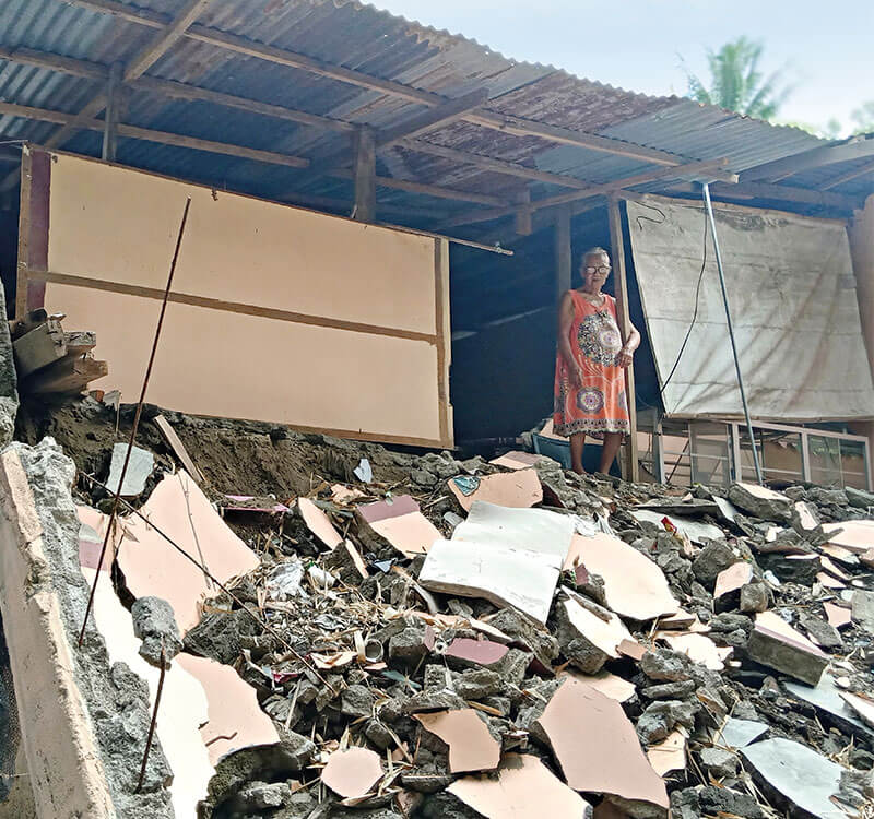 Teresita standing in her home, with the floor caved in and covered with debris beneath her